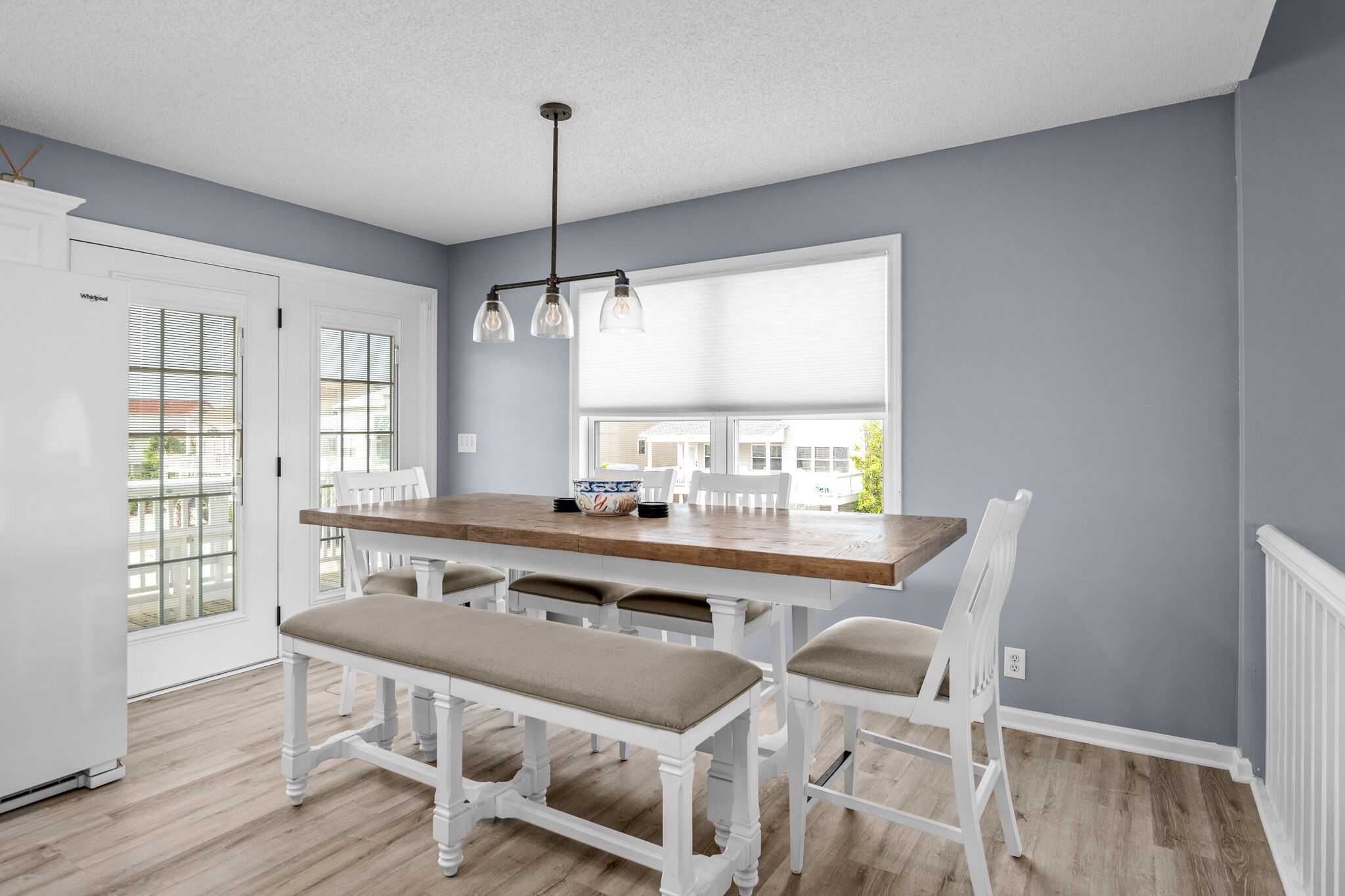 Een eethoek met een houten tafel en witte stoelen, een hanglamp en uitzicht door een raam.