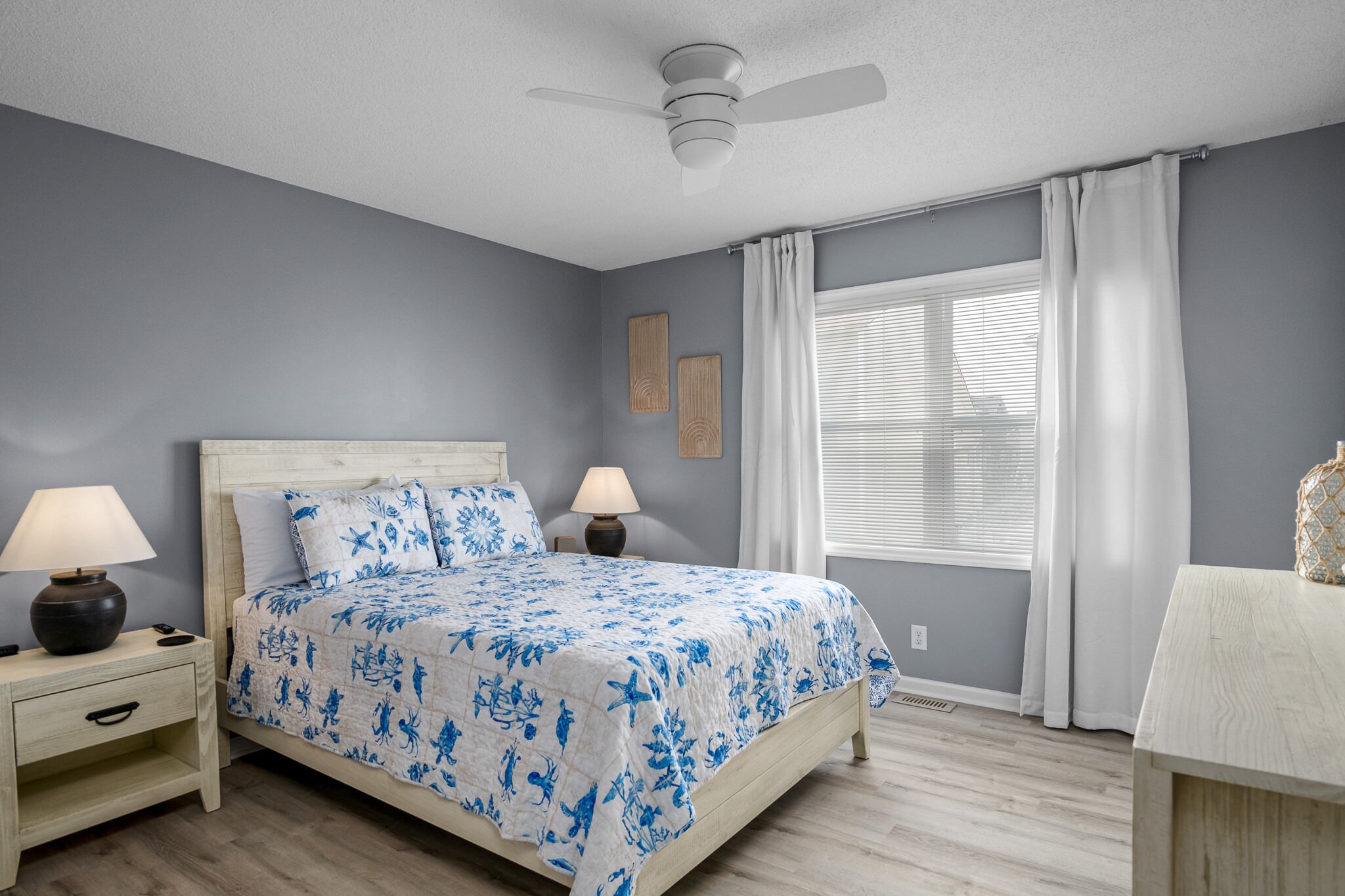A bedroom with a bed, two bedside tables, a ceiling fan, a window with curtains, and a wall-mounted artwork.