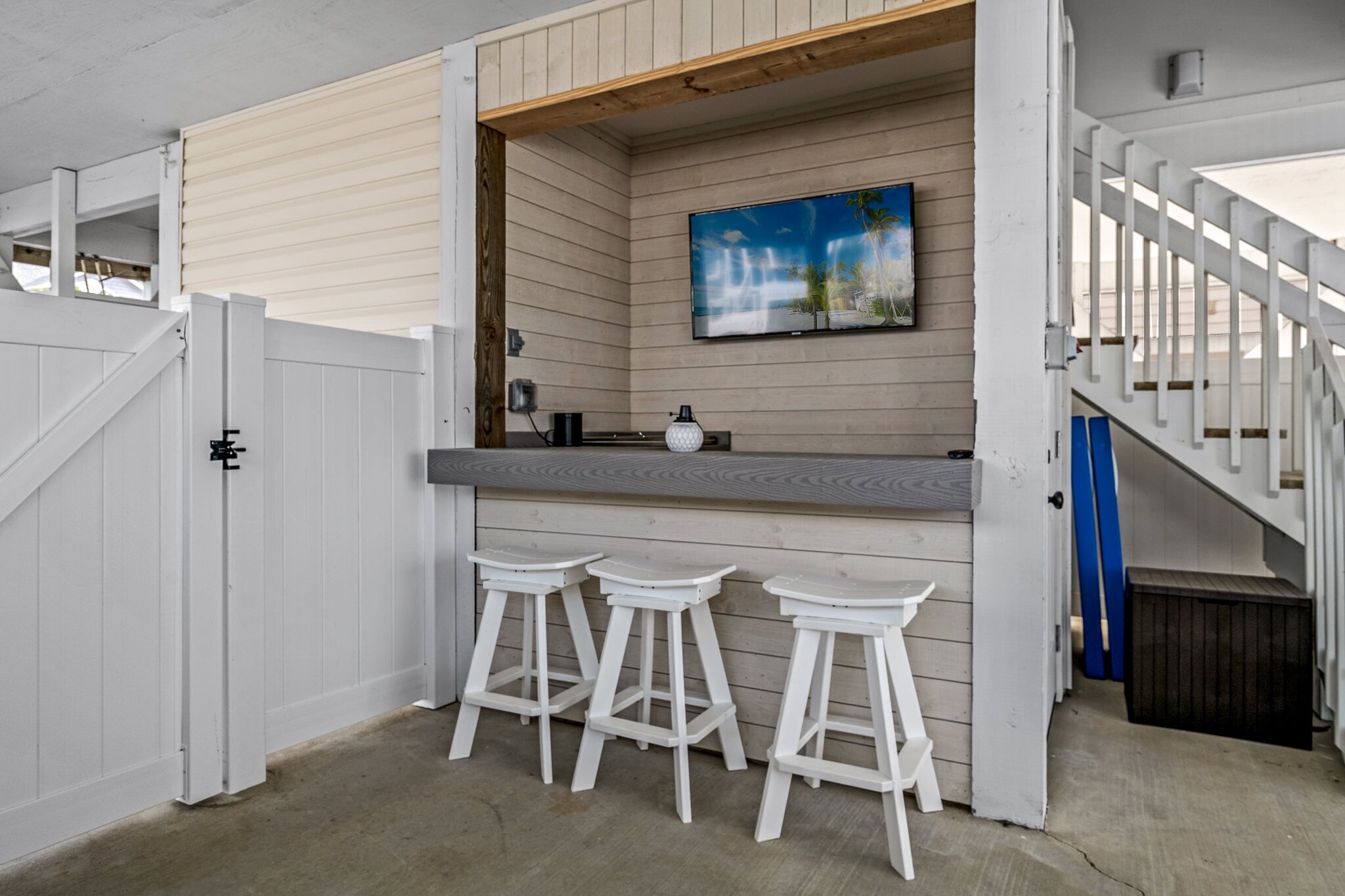 A bar area with a wooden counter, three white stools, and a painting of a tropical scene.