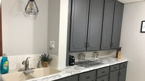 A kitchen with a marble countertop, grey cabinets, a sink, a coffee maker, and a dish rack. - 1 Block to the Beach! Firepit, 2 Bikes, Sleeps 5 (Two Rivers)