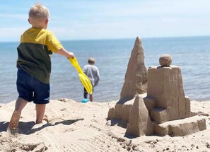 Ein Kind spielt am Strand mit Sand, im Hintergrund ein großes Sandkastenschloss und ruhige See.