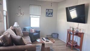 A living room with a brown leather sofa, a blue armchair, a wooden coffee table, a television, and a wooden side table with board games. - 1 Block to the Beach! Firepit, 2 Bikes, Sleeps 5 (Two Rivers)