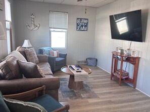 A living room with a brown leather sofa, a blue armchair, a wooden coffee table, a television, and a wooden side table with board games. - 1 Block to the Beach! Firepit, 2 Bikes, Sleeps 4 (Two Rivers)
