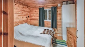 Puuinen mökkihuone, jossa on sänky, lipasto ja ikkuna, jossa on verhot.
