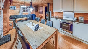 Tunnelmallinen mökin sisustus, jossa on puinen ruokapöytä, tuoleja, sohva, liesi ja keittiö sekä ruokailutila, jonka yllä on koristeellinen kattokruunu.