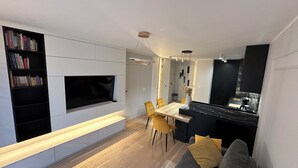 A modern living room with a flat-screen TV, a bookshelf, a dining area with yellow chairs, and a kitchen area with a black countertop. - Luxury Apartment Near Paris (Issy-les-Moulineaux)