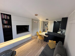 Ein modernes Wohnzimmer mit Fernseher, Bücherregal, Essbereich mit gelben Stühlen und Küchenzeile mit schwarzer Arbeitsplatte.