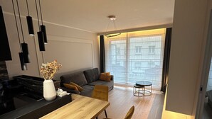 A modern kitchen with a central island, a dining area with wooden chairs, a sofa, and a view of the city through large windows. - Luxury Apartment Near Paris (Issy-les-Moulineaux)