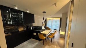A modern kitchen with dark cabinetry, a marble backsplash, and a central island. There is a dining area with a wooden table and yellow chairs. - Luxury Apartment Near Paris (Issy-les-Moulineaux)