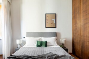 Ein ordentlich eingerichtetes Hotelzimmer mit einem Bett, zwei Nachttischlampen, einem gerahmten Kunstwerk an der Wand und einem Holzschrank.