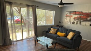 Ein Wohnzimmer mit Sofa, Couchtisch und Sesseln. Es gibt ein großes Fenster mit Vorhängen, einen Deckenventilator und ein Bild an der Wand.