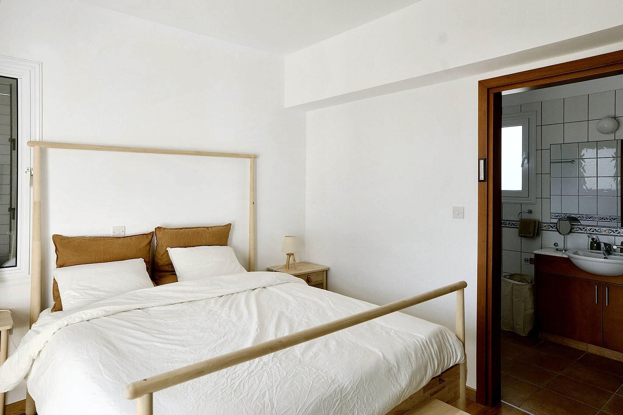 A bedroom with a wooden bed frame, white bedding, and a nightstand with a lamp.