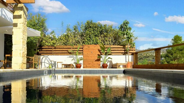 A pool with a clear reflection, surrounded by wooden deck and potted plants, with a stone pillar and outdoor furniture. - Romantic High-End Pool 3-bedroom villa in Peyia, Cyprus (Peyia)