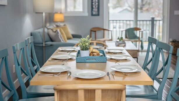 Un coin repas avec une table en bois dressée pour six personnes, des chaises bleues, une petite plante en pot et un plateau bleu contenant de la nourriture.