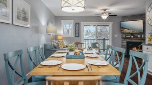 A dining area with a wooden table set for four, blue chairs, a chandelier, and a television mounted on the wall.