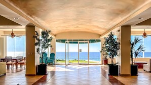 A spacious indoor area with a view of the ocean, featuring a dining area with chairs and a table, large potted plants, and a ceiling fan.