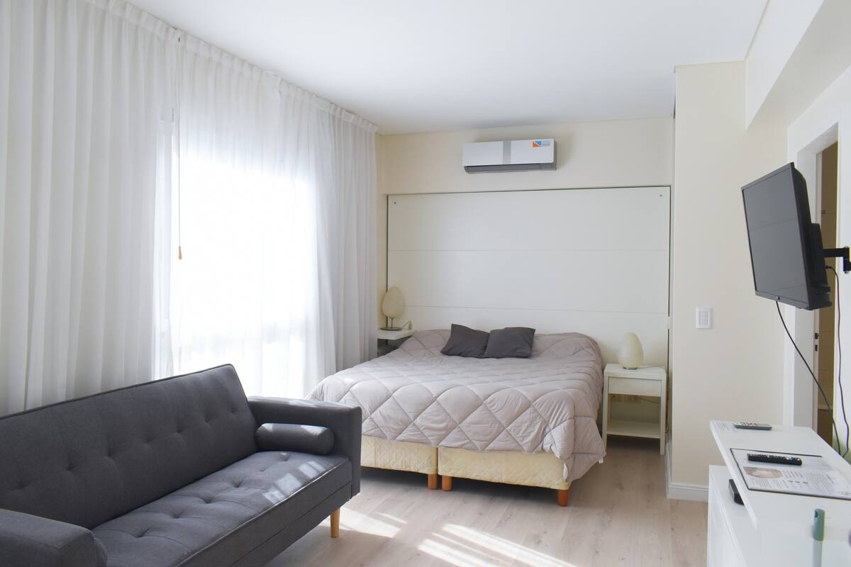 A modern hotel room with a bed, a sofa, a TV, and an air conditioning unit.