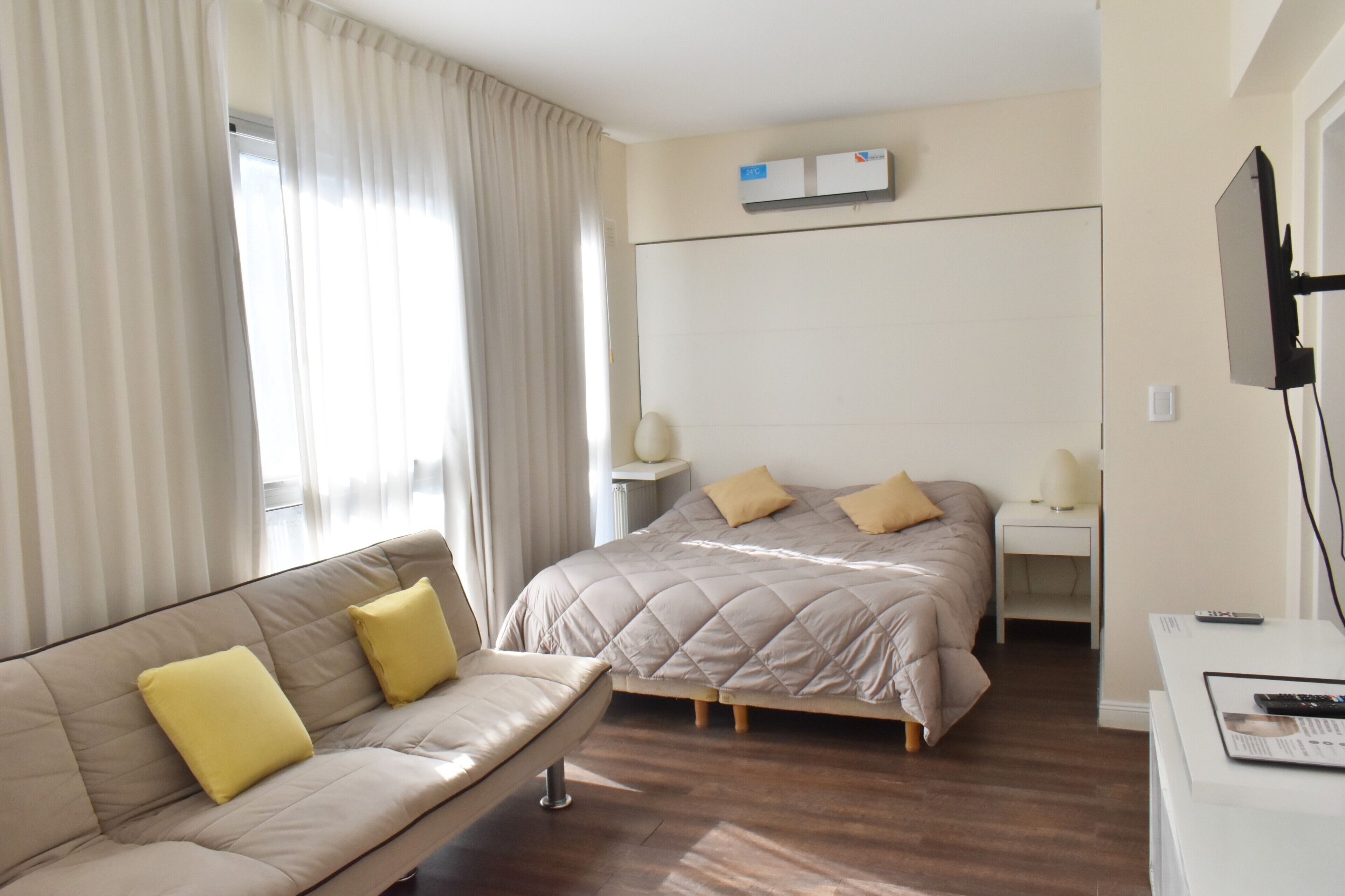 A hotel room with a bed, a sofa, a bedside table, a TV, and a window with curtains.