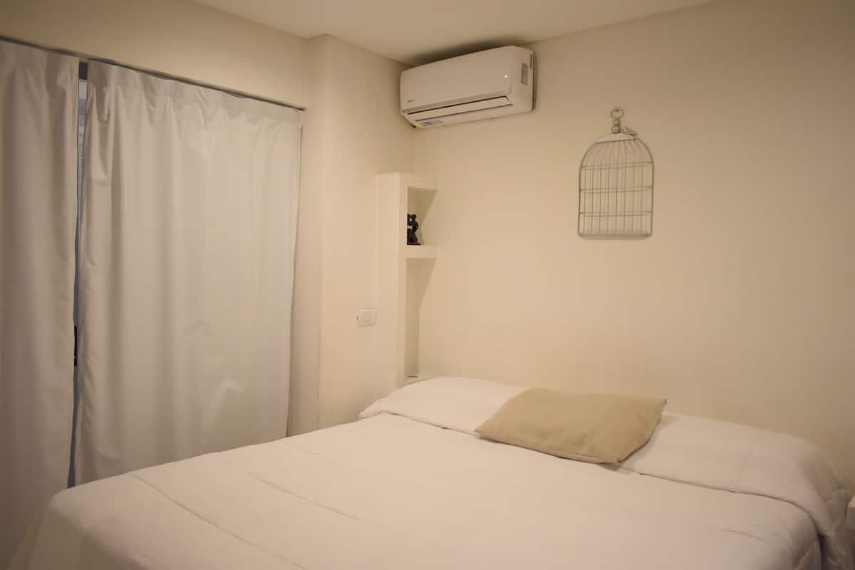 A bedroom with a bed, white curtains, an air conditioning unit, a shelf with a decorative item, and a wall-mounted birdcage.