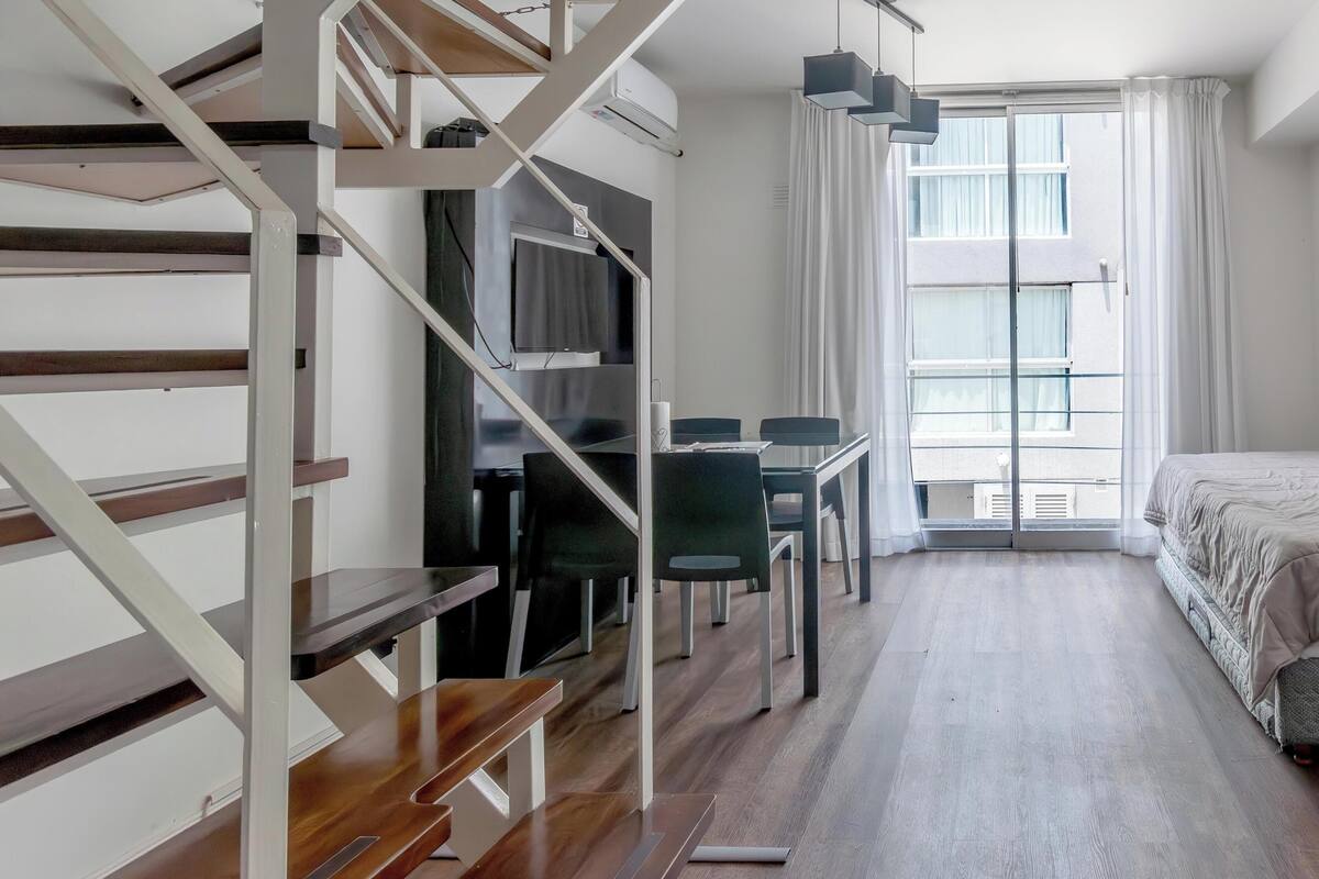 A modern apartment with a spiral staircase, a dining area, a bed, and a large window.