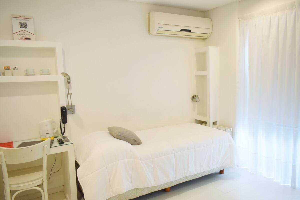 A hotel room with a bed, a desk with a chair, a telephone, and an air conditioning unit.