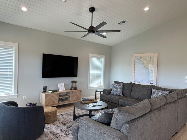 A modern living room with a grey sofa, a coffee table, a TV mounted on the wall, and a ceiling fan. - Beautiful home on the river, mins to the Gulf; fire-pit, bar, view (Steinhatchee)