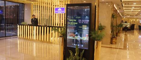 Zona de recepción de hotel con un menú digital, un mostrador de recepción y una recepcionista.
