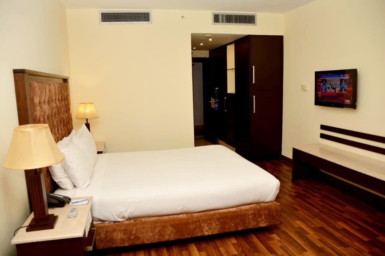 A hotel room with a bed, a nightstand with a lamp, a TV, and a wooden floor.
