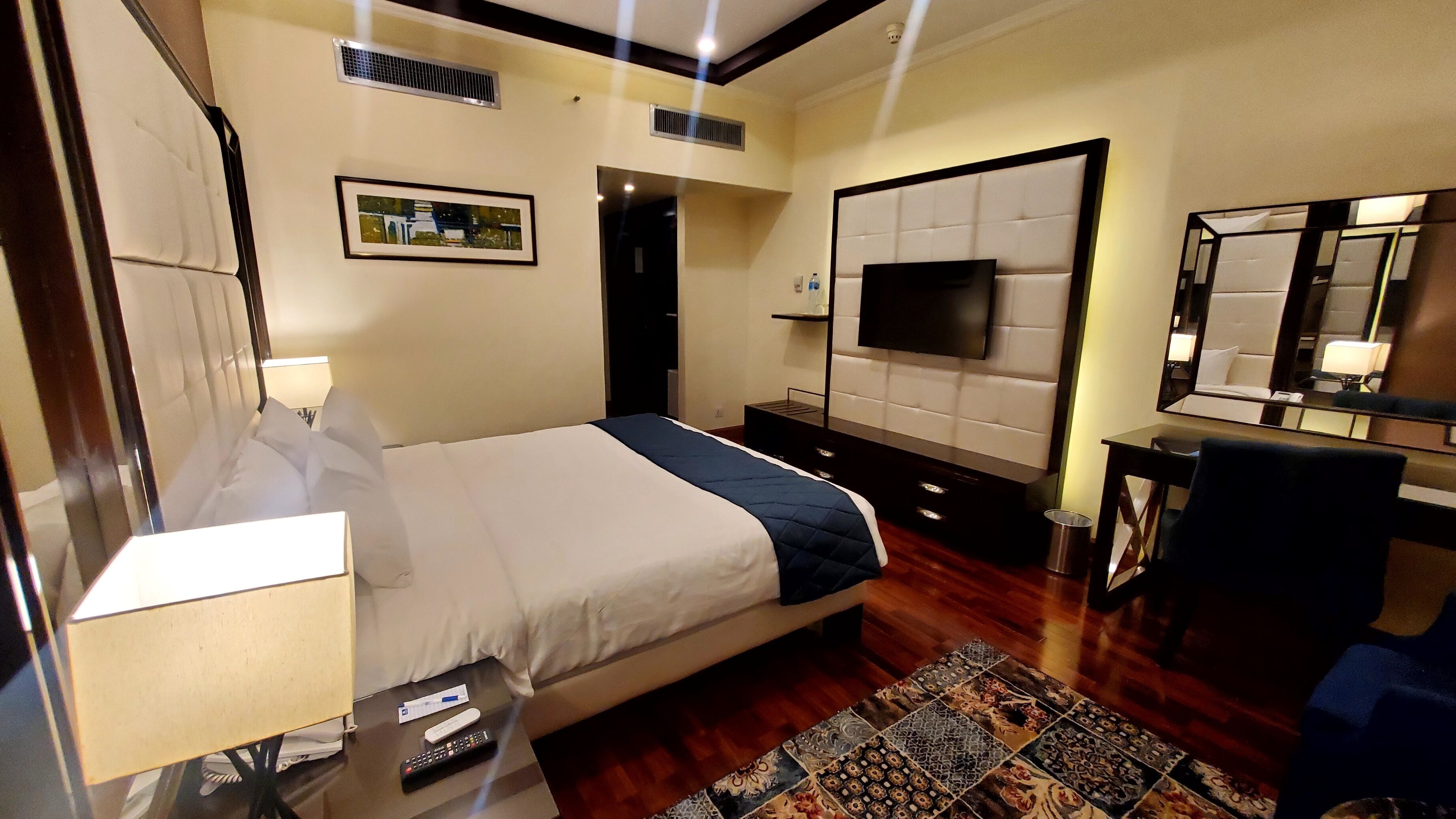 A hotel room with a bed, bedside table, lamp, TV, and a mirror.