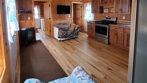A cabin interior with a wooden floor, a sofa, a TV, a kitchen area, and a bed with a patterned blanket.
