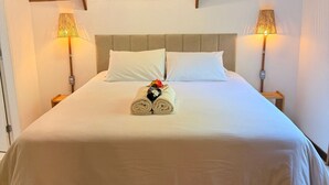 Una cama bien hecha con edredón blanco, dos almohadas y una toalla enrollada con un llavero. Hay dos lámparas de noche con pantallas a cada lado de la cama.