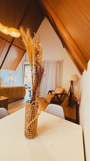 Une chambre à coucher confortable, aménagée dans un espace en mezzanine avec un plafond en pente, une grande fenêtre et un coin repas agrémenté d’un vase en osier posé sur la table.