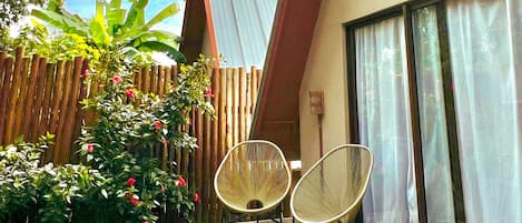 Una casa moderna con techo inclinado, una terraza de madera, dos sillas de mimbre y un amplio ventanal con cortinas.