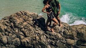 Una costa rocosa con aguas turquesas cristalinas, una montaña distante y una playa al fondo.