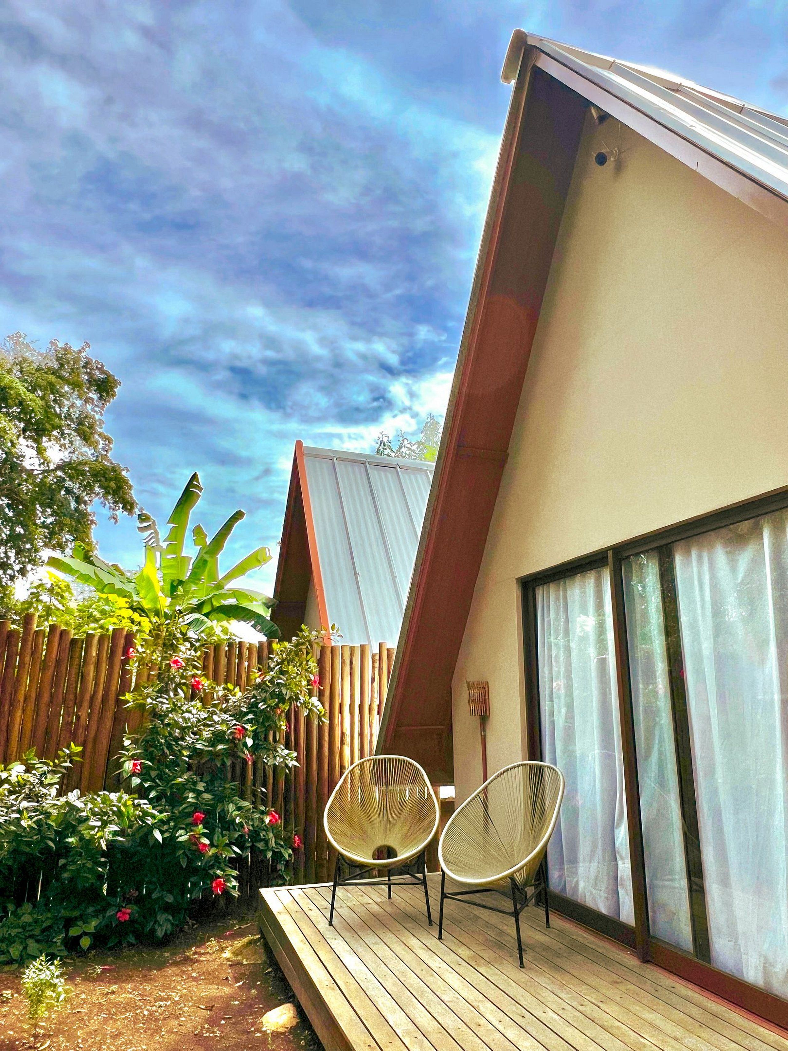Una casa moderna con techo inclinado, una terraza de madera, dos sillas de mimbre y un amplio ventanal con cortinas.
