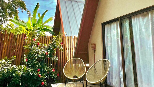 Una casa moderna con techo inclinado, una terraza de madera, dos sillas de mimbre y un amplio ventanal con cortinas.