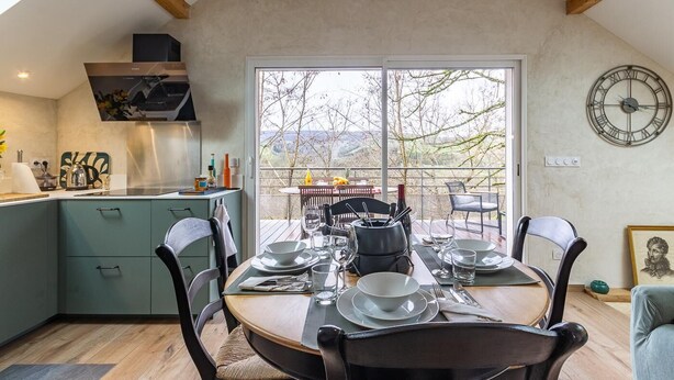 Un comedor con una mesa dispuesta para cuatro personas, una cocina con una estufa moderna, y un ventanal con vistas a árboles y colinas.
