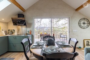 A dining area with a table set for four, a kitchen with a modern stove, and a large window offering a view of trees and hills. - L'ecrin - Saint Paul SUR Yenne (SAINT PAUL SUR YENNE)