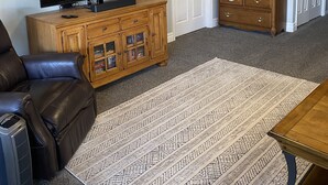 Sala de estar com televisão de tela plana em móvel de madeira, poltrona de couro, tapete estampado e mesa de madeira.