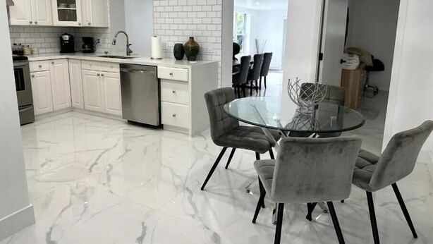 Una cocina moderna con armarios blancos, lavavajillas y piso de mármol. Cuenta con un comedor con mesa de vidrio y sillas grises.