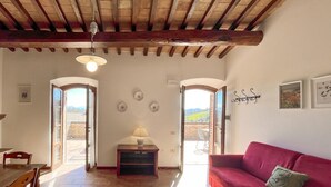 Ein großzügiger Wohnbereich mit einem roten Sofa, einem Holzesstisch und einer Lampe. Zwei Türen führen zu einem Balkon mit Blick auf die Landschaft.