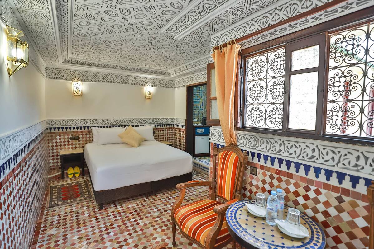 A room with a bed, a chair, a small table with glasses and bottles, and a window with decorative ironwork.