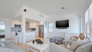 A modern living room with a sofa, a coffee table, and a kitchen area with white cabinets and a flat-screen TV. - 5951- South Of Stars - South Nags Head Escape w/ Pool & Hot Tub (Nags Head)