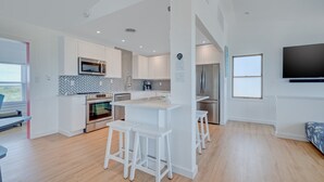 A modern kitchen with white cabinetry, a microwave, an oven, and a refrigerator. There are three white stools around a central island. A flat-screen TV is mounted on the wall. - 5951- South Of Stars - South Nags Head Escape w/ Pool & Hot Tub (Nags Head)