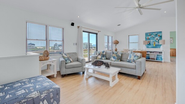 A spacious living room with a white ceiling fan, two grey sofas with patterned cushions, a white coffee table, and a view of the city through large windows. - 5951- South Of Stars - South Nags Head Escape w/ Pool & Hot Tub (Nags Head)