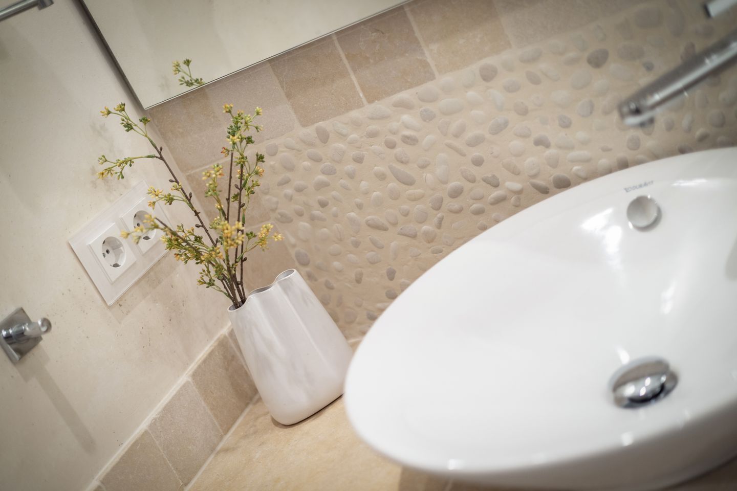 Une salle de bain avec un lavabo blanc, des carreaux beiges imitant des galets et un vase blanc contenant des fleurs jaunes.