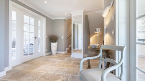 A spacious hallway with stone flooring, a white chair, and a lamp on a table. - Wattblick. Reet. Sauna. - Hideaways (Sylt)
