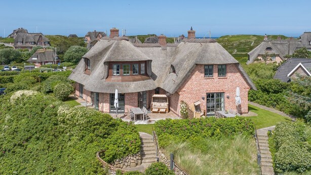A large, two-story house with a thatched roof, surrounded by lush greenery and a clear view of the sea. - Wattblick. Reet. Sauna. - Hideaways (Sylt)