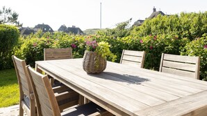 Une table à manger en bois, accompagnée de quatre chaises, un panier tressé rempli de fleurs, et en arrière-plan, une végétation luxuriante et des maisons.