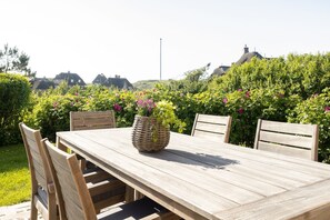 A wooden dining table with four chairs, a woven basket with flowers, and a backdrop of lush greenery and houses.